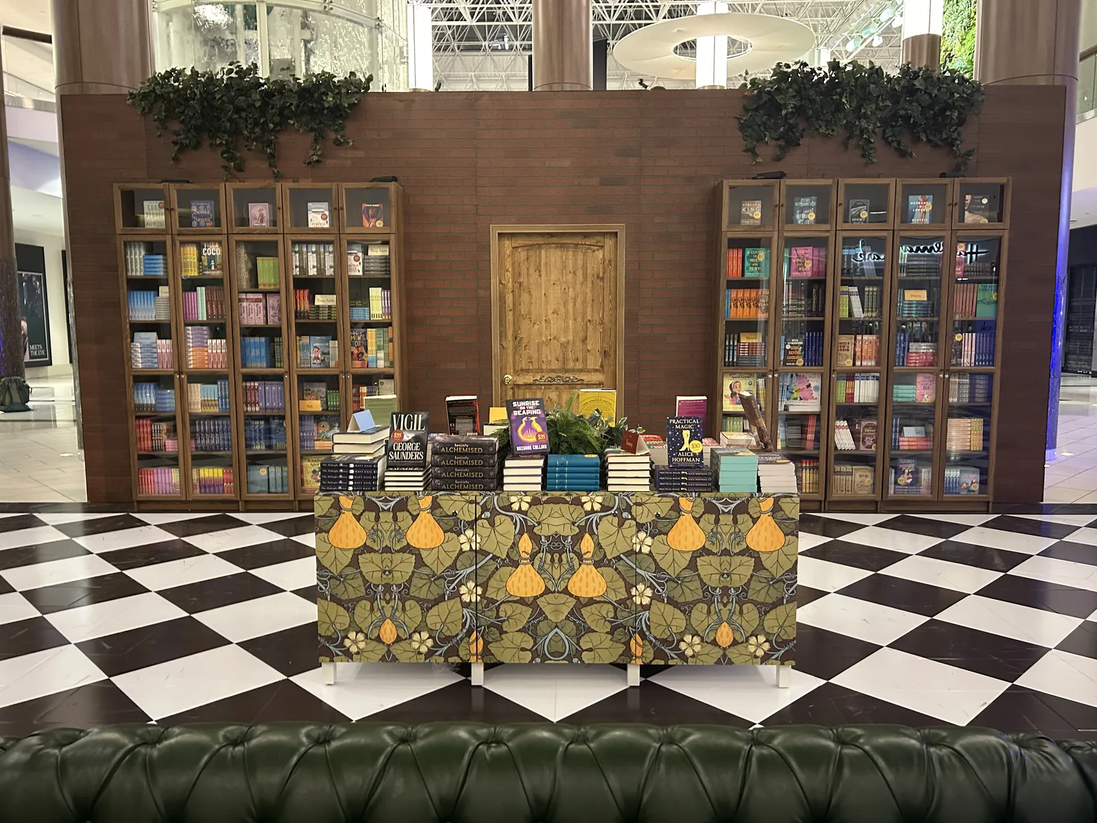 The Book Corner bookcases on a brick wall around a wooden door, with a patterned book table on the checkerboard floor
