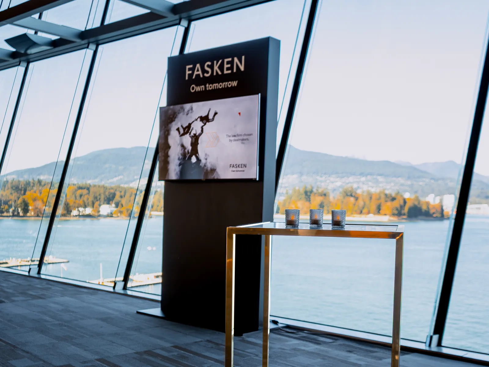 Fasken branded TV wall beside a gold high-top table in the Ocean View Foyer, with Vancouver Harbour behind the windows