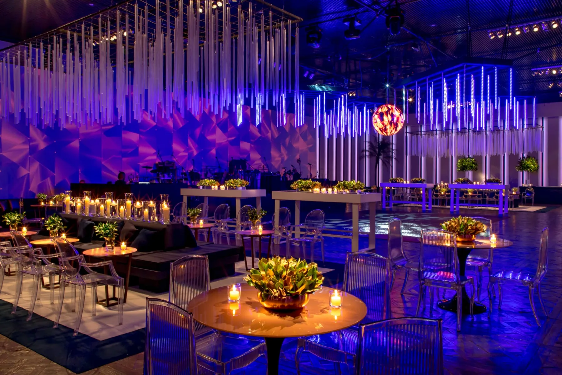 Ballroom in blue and violet light with hanging acrylic rods, a suspended mirror sphere, candlelit tables and clear acrylic chairs